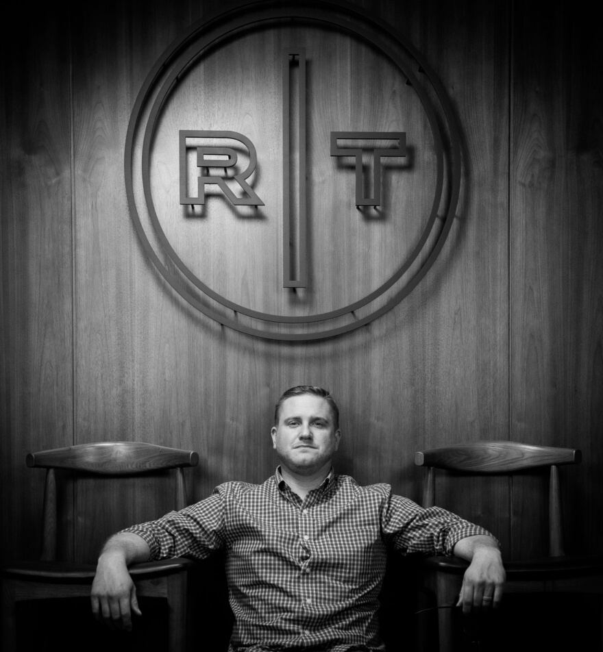 Round Table Studios employee sitting by chairs on the ground with Round Table Studios Logo behind him.